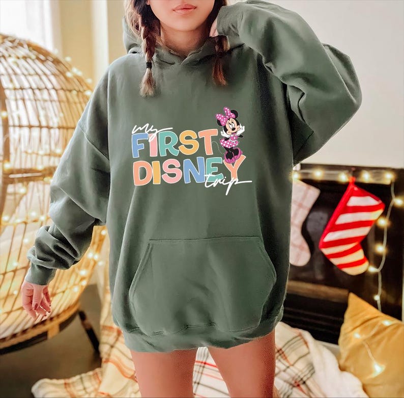 May include: An olive green hooded sweatshirt with a front pocket. The sweatshirt features the text "My First Disney Trip" in colorful letters, along with a graphic of Minnie Mouse. The sweatshirt has long sleeves and a drawstring hood.
