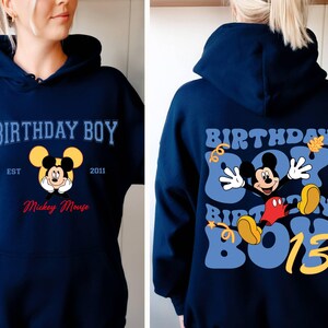 K&ouml;nnte beinhalten: Ein marineblauer Hoodie mit einem Mickey-Mouse-Motiv und dem Text "Birthday Boy Est 2011 Mickey Mouse" auf der Vorderseite. Die R&uuml;ckseite des Hoodies zeigt ein gr&ouml;&szlig;eres Mickey-Mouse-Motiv mit dem Text "Birthday Boy 13" in einer Retro-Schrift.