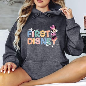 May include: Dark gray hooded sweatshirt with the text "My First Disney Trip" in colorful letters. A Minnie Mouse graphic is on the right side. The sweatshirt has a drawstring hood and a front pocket.