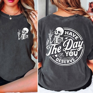 May include: A dark gray t-shirt with a white skeleton graphic on the front and a white circle graphic on the back that says "Have The Day You Deserve".