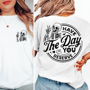 May include: White t-shirt with a black skeleton graphic on the front and a black graphic on the back that says "Have the Day You Deserve" with a skeleton graphic.