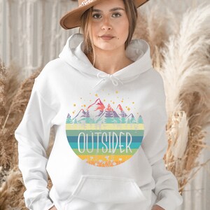 May include: White hoodie with a colorful mountain and forest graphic. The word "OUTSIDER" is printed in a bold font across the design. The hoodie has a drawstring hood and a front pocket.