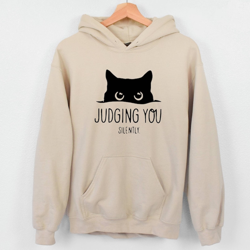 K&ouml;nnte beinhalten: Ein beiger Hoodie mit einer schwarzen Katze, die &uuml;ber den Rand schaut, und dem Text "JUDGING YOU SILENTLY."