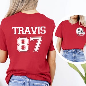 May include: Red t-shirt with white text on the back that says "TRAVIS 87". The front of the shirt has a white football helmet with the text "87 TRAVIS" on it.
