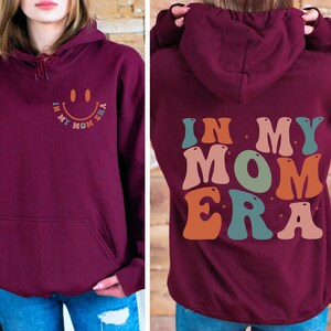 May include: Burgundy hoodie with a retro-style graphic that says "In My Mom Era" in colorful letters. The graphic features a smiley face with a star on each side.