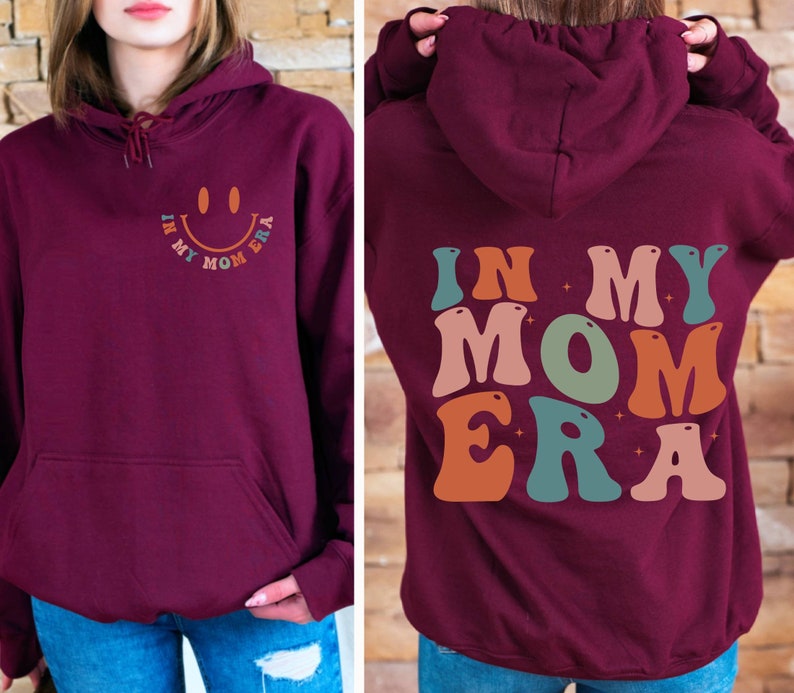 May include: Burgundy hoodie with a retro-style graphic that says "In My Mom Era" in colorful letters. The graphic features a smiley face with a star on each side.