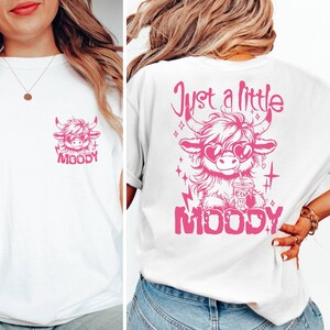 Puede incluir: Camiseta blanca con un gr&aacute;fico rosa de una vaca de las tierras altas de dibujos animados con gafas de sol y sosteniendo una bebida. El texto "Just a little MOODY" est&aacute; impreso sobre la vaca y "MOODY" est&aacute; impreso debajo de la vaca.