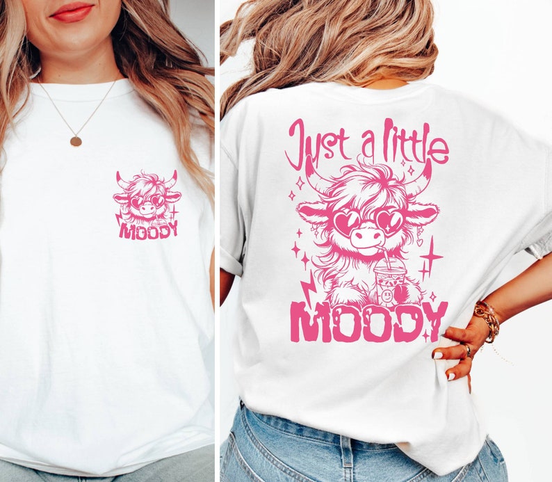 Puede incluir: Camiseta blanca con un gr&aacute;fico rosa de una vaca de las tierras altas de dibujos animados con gafas de sol y sosteniendo una bebida. El texto "Just a little MOODY" est&aacute; impreso sobre la vaca y "MOODY" est&aacute; impreso debajo de la vaca.