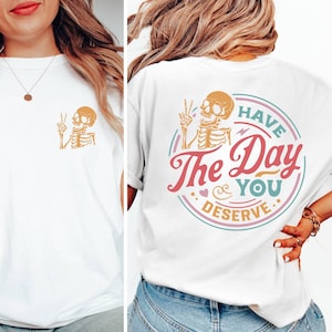 May include: White t-shirt with a skeleton graphic on the front and a graphic on the back that says "Have the Day You Deserve" with a skeleton graphic.