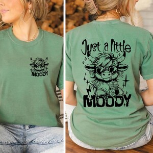 Puede incluir: Una camiseta verde con un gr&aacute;fico negro de una vaca de las tierras altas de dibujos animados que lleva gafas de sol y bebe una bebida. El texto "Just a little moody" est&aacute; por encima de la vaca y la palabra "MOODY" est&aacute; por debajo de la vaca.