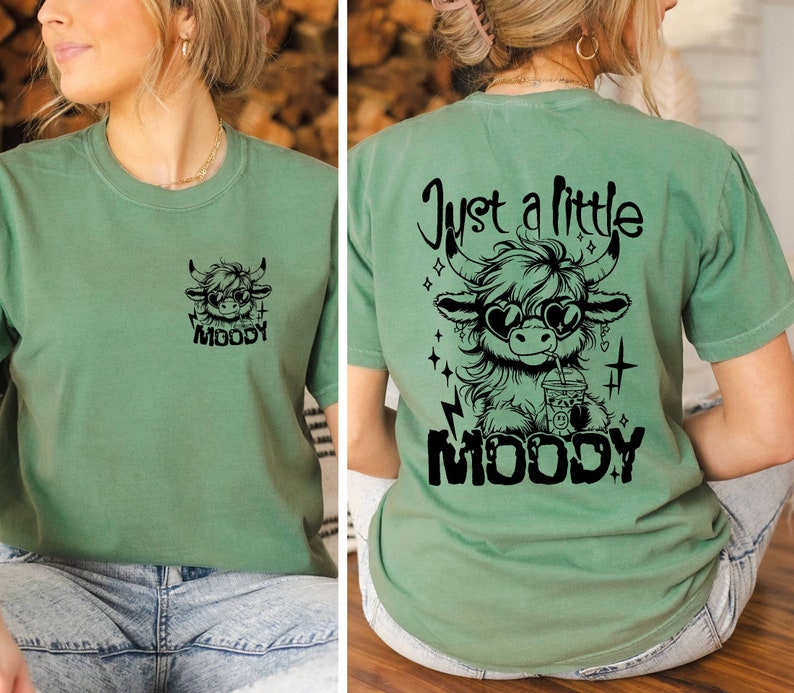 Puede incluir: Una camiseta verde con un gr&aacute;fico negro de una vaca de las tierras altas de dibujos animados que lleva gafas de sol y bebe una bebida. El texto "Just a little moody" est&aacute; por encima de la vaca y la palabra "MOODY" est&aacute; por debajo de la vaca.