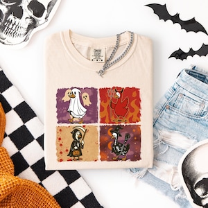 May include: Cream-colored t-shirt featuring a Halloween-themed graphic with four panels. Each panel depicts a cartoon duck in a costume: ghost, devil, grim reaper, and witch. The shirt is accessorized with a silver chain.