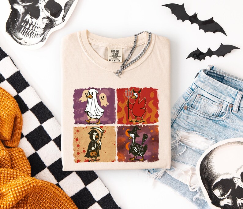 May include: Cream-colored t-shirt featuring a Halloween-themed graphic with four panels. Each panel depicts a cartoon duck in a costume: ghost, devil, grim reaper, and witch. The shirt is accessorized with a silver chain.