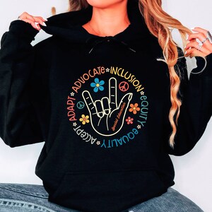 May include: Black hoodie with a graphic print of a hand making the American Sign Language sign for "I love you". The graphic is surrounded by a circle with the words "Adapt, Accept, Advocate, Inclusion, Equity, Equality" in a rainbow color scheme.