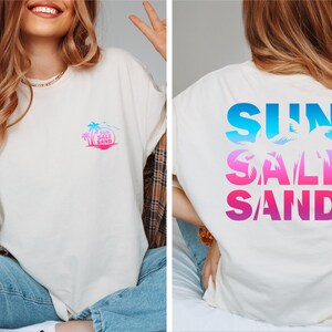 Pode incluir: Camiseta creme com um design tropical. A frente apresenta um pequeno gr&aacute;fico colorido de uma palmeira e as palavras "Sun Salt Sand". A parte de tr&aacute;s da camisa exibe as palavras "SUN SALT SAND" em letras grandes gradientes azuis e rosa.