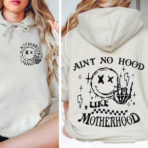 May include: A light beige hoodie with a black graphic print on the front and back. The graphic on the front says "Motherhood" with a smiley face and a hand with a middle finger extended. The graphic on the back says "Ain't No Hood Like Motherhood" with a smiley face and a skeleton hand with a middle finger extended.