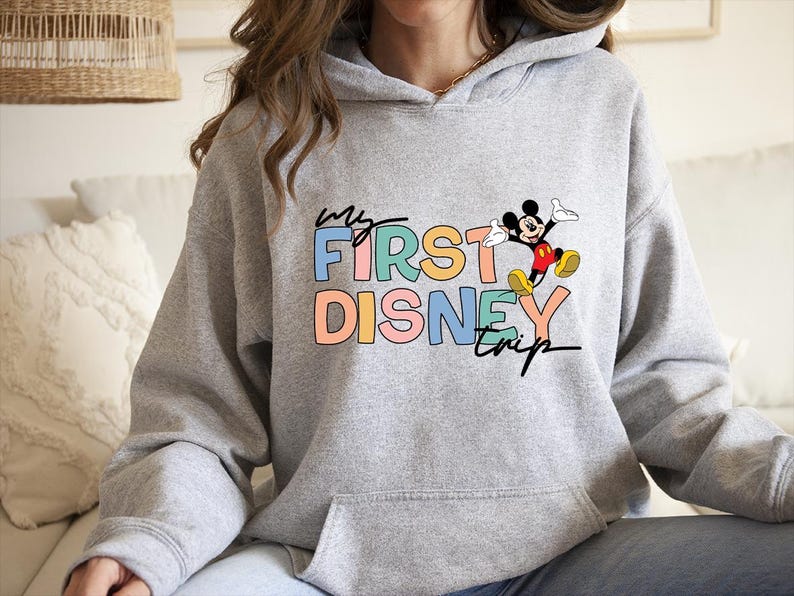 May include: A light gray hoodie with a front graphic that reads "my FIRST DISNEY trip" in colorful block letters. A cartoon Mickey Mouse is jumping next to the text. The hoodie has a front pocket and a hood.