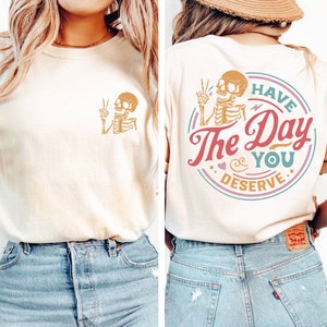 May include: A light beige t-shirt with a skeleton graphic on the front and a circular graphic on the back that says "Have the Day You Deserve" with a skeleton graphic inside the circle.