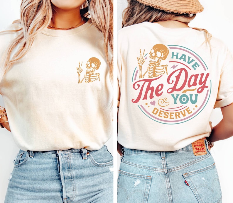 May include: A light beige t-shirt with a skeleton graphic on the front and a circular graphic on the back that says "Have the Day You Deserve" with a skeleton graphic inside the circle.