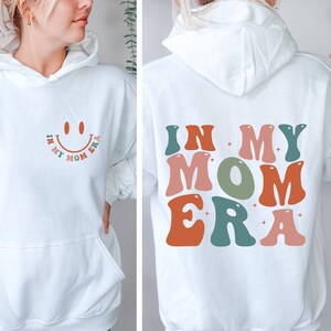May include: White hoodie with a smiley face and the text "In My Mom Era" on the front and the text "In My Mom Era" in colorful retro letters on the back.