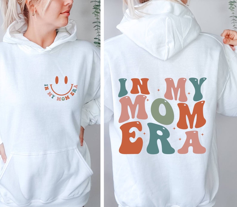 May include: White hoodie with a smiley face and the text "In My Mom Era" on the front and the text "In My Mom Era" in colorful retro letters on the back.