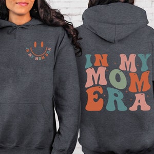 May include: A dark gray hoodie with the text "In My Mom Era" in colorful retro letters on the back. The front of the hoodie has a smiley face with the same text in a smaller font.