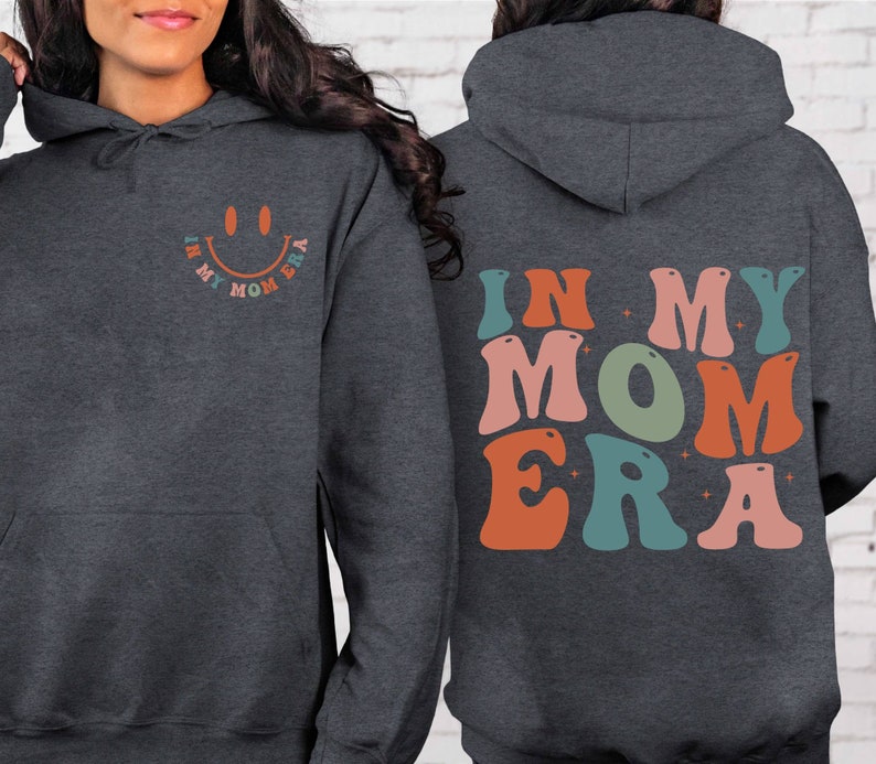 May include: A dark gray hoodie with the text "In My Mom Era" in colorful retro letters on the back. The front of the hoodie has a smiley face with the same text in a smaller font.