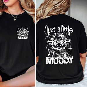 Puede incluir: Camiseta negra con un gr&aacute;fico blanco de una vaca de las tierras altas de dibujos animados con gafas de sol y sosteniendo una taza. El texto "Just a little MOODY" est&aacute; impreso en la camiseta.