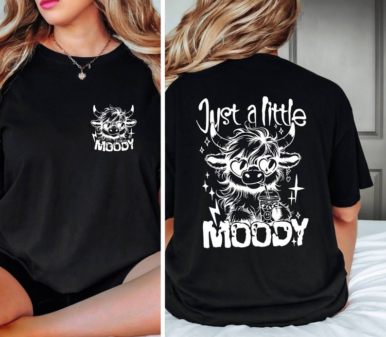 Puede incluir: Camiseta negra con un gr&aacute;fico blanco de una vaca de las tierras altas de dibujos animados con gafas de sol y sosteniendo una taza. El texto "Just a little MOODY" est&aacute; impreso en la camiseta.