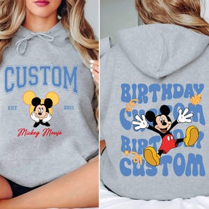 K&ouml;nnte beinhalten: Grauer Kapuzenpullover mit einem blauen und wei&szlig;en Aufdruck von Mickey Mouse. Der Aufdruck lautet "Custom" mit "Est 2011" und "Mickey Mouse" darunter. Die R&uuml;ckseite des Pullovers hat einen blauen und wei&szlig;en Aufdruck, der "Birthday Custom Birthday Custom" lautet.