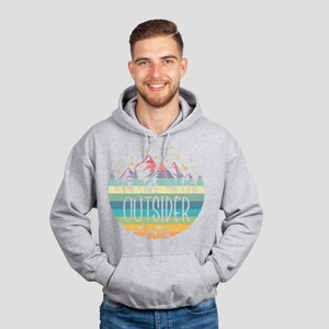 May include: A light gray hoodie with a colorful graphic of mountains, trees, and the word "OUTSIDER" in a retro style. The hoodie has a drawstring hood and a front pocket.