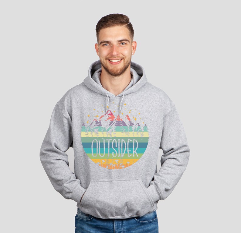 May include: A light gray hoodie with a colorful graphic of mountains, trees, and the word "OUTSIDER" in a retro style. The hoodie has a drawstring hood and a front pocket.