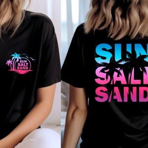 Pode incluir: Camiseta preta com um design colorido de palmeiras e as palavras "SUN SALT SAND" em gradiente de azul, rosa e roxo. O design est&aacute; na frente e atr&aacute;s, evocando um tema de praia.