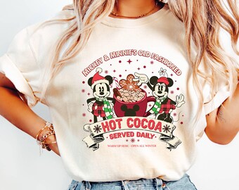 May include: Cream-colored t-shirt featuring a graphic of Mickey and Minnie Mouse with a hot cocoa theme. The design includes the text "Mickey & Minnie's Old Fashioned Hot Cocoa Served Daily" with snowflakes and a banner. The characters are wearing holiday attire.