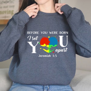 Puede incluir: Una sudadera azul oscuro con un dise&ntilde;o gr&aacute;fico blanco que presenta un coraz&oacute;n hecho de piezas de rompecabezas en rojo, amarillo, azul y verde. El texto dice "Antes de que nacieras, te apart&eacute; Jerem&iacute;as 1:5".