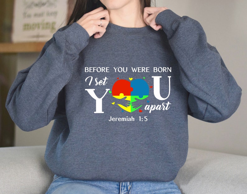 Puede incluir: Una sudadera azul oscuro con un dise&ntilde;o gr&aacute;fico blanco que presenta un coraz&oacute;n hecho de piezas de rompecabezas en rojo, amarillo, azul y verde. El texto dice "Antes de que nacieras, te apart&eacute; Jerem&iacute;as 1:5".
