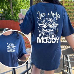 Puede incluir: Una camiseta azul marino con un gr&aacute;fico blanco de una vaca de las tierras altas con gafas de sol y sosteniendo una taza. El texto "Just a little MOODY" est&aacute; impreso debajo de la vaca.