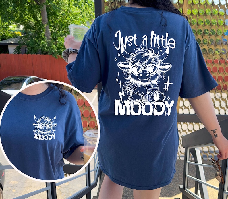 Puede incluir: Una camiseta azul marino con un gr&aacute;fico blanco de una vaca de las tierras altas con gafas de sol y sosteniendo una taza. El texto "Just a little MOODY" est&aacute; impreso debajo de la vaca.