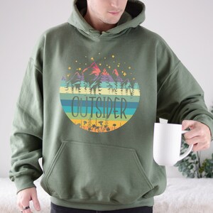 May include: Olive green hoodie with a colorful mountain and forest graphic. The graphic includes the word "OUTSIDER" in a circular design with horizontal stripes. The hoodie has a front pocket and a drawstring hood.