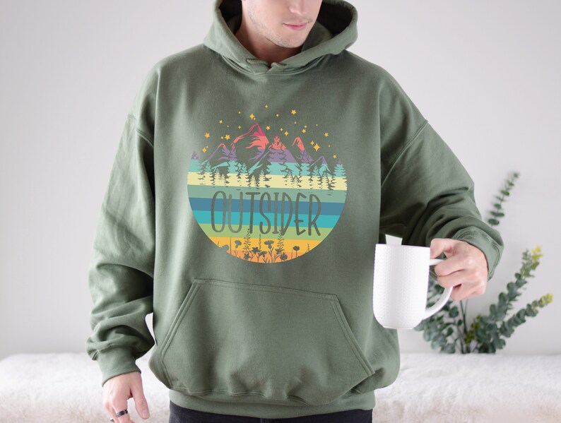 May include: Olive green hoodie with a colorful mountain and forest graphic. The graphic includes the word "OUTSIDER" in a circular design with horizontal stripes. The hoodie has a front pocket and a drawstring hood.