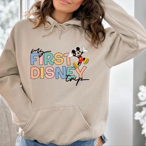 May include: Beige hoodie with a front pocket and a drawstring hood. The front features the text "My First Disney Trip" in colorful block letters, with a cartoon Mickey Mouse graphic.