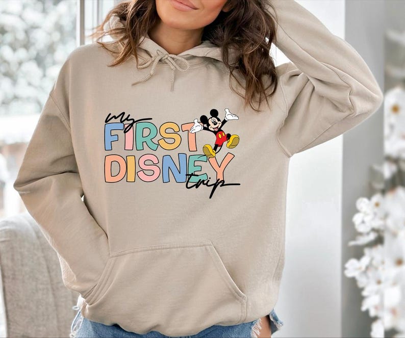 May include: Beige hoodie with a front pocket and a drawstring hood. The front features the text "My First Disney Trip" in colorful block letters, with a cartoon Mickey Mouse graphic.