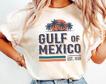 May include: A cream colored t-shirt with a vintage-style graphic of palm trees and a sunset. The text "GULF OF MEXICO EST. 1550" is printed on the shirt.