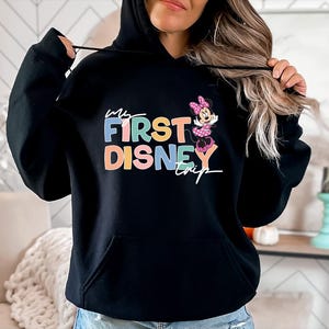 May include: Black hooded sweatshirt with the text "My First Disney Trip" in colorful letters, accompanied by a Minnie Mouse graphic. The sweatshirt has a front pocket and a drawstring hood.
