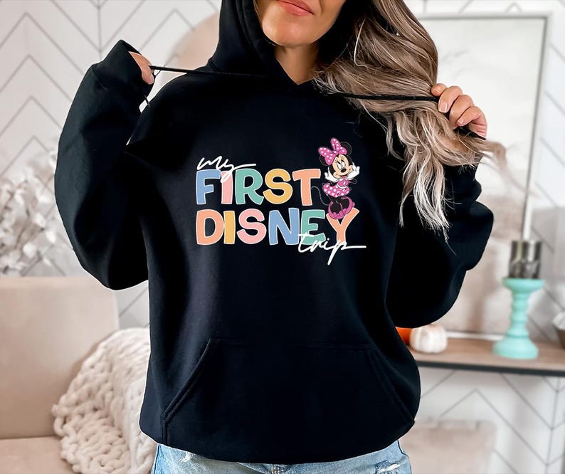 May include: Black hooded sweatshirt with the text "My First Disney Trip" in colorful letters, accompanied by a Minnie Mouse graphic. The sweatshirt has a front pocket and a drawstring hood.