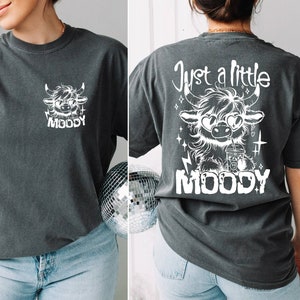 Puede incluir: Una camiseta gris oscuro con un gr&aacute;fico blanco de una vaca de las tierras altas de dibujos animados con gafas de sol y sosteniendo una bebida. El texto "Just a little MOODY" est&aacute; impreso en la parte posterior de la camiseta.
