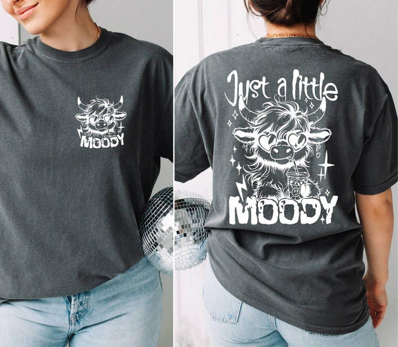 Puede incluir: Una camiseta gris oscuro con un gr&aacute;fico blanco de una vaca de las tierras altas de dibujos animados con gafas de sol y sosteniendo una bebida. El texto "Just a little MOODY" est&aacute; impreso en la parte posterior de la camiseta.