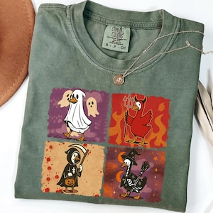 May include: Green Comfort Colors t-shirt featuring four Halloween-themed duck illustrations. The ducks are dressed as a ghost, devil, grim reaper, and witch. A gold necklace is draped across the shirt.