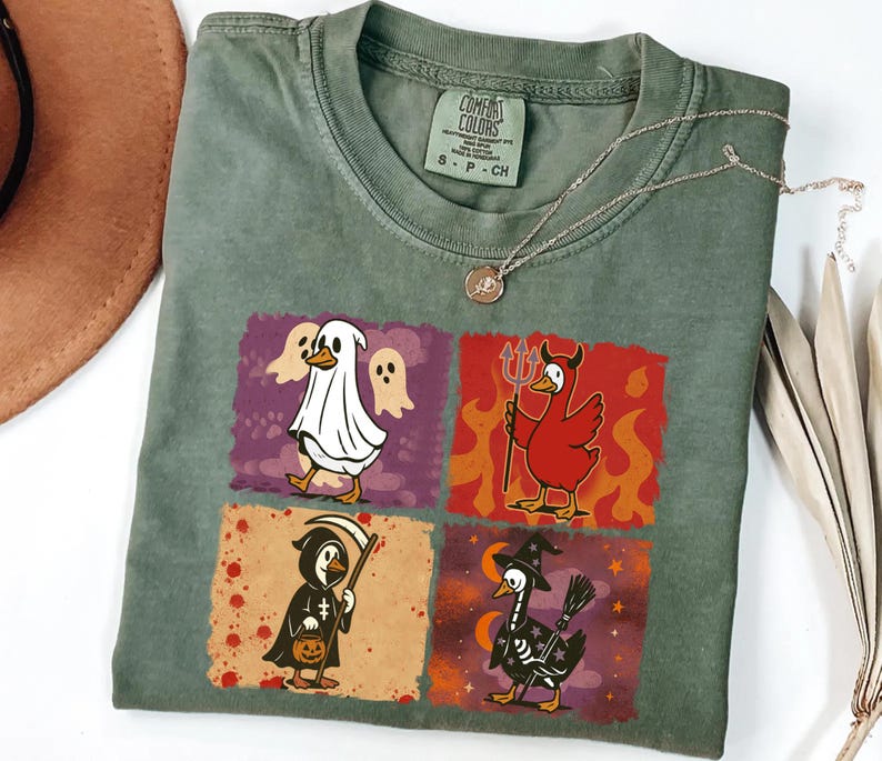 May include: Green Comfort Colors t-shirt featuring four Halloween-themed duck illustrations. The ducks are dressed as a ghost, devil, grim reaper, and witch. A gold necklace is draped across the shirt.