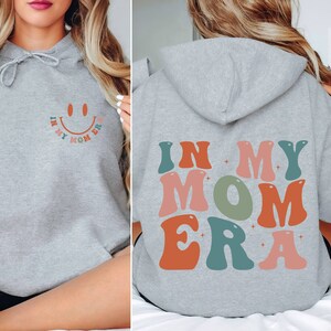 May include: Gray hooded sweatshirt with the text "In My Mom Era" in colorful retro font on the back. The front of the sweatshirt has a smiley face with the text "In My Mom Era" around it.